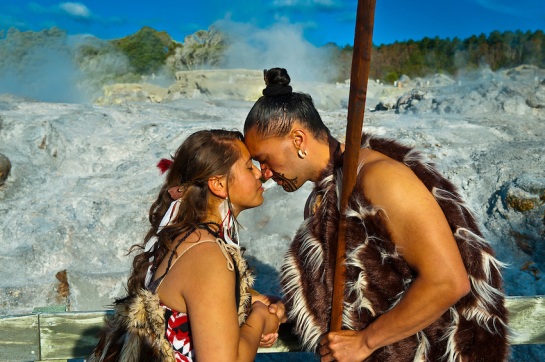 A Maori man with ta moko (facial tattoo) and woman doing hongi (traditional Maori greeting) with the Pohutu Geyser behind, Te Puia (New Zealand Maori Arts &amp; Crafts Institute), Rotorua, New Zealand